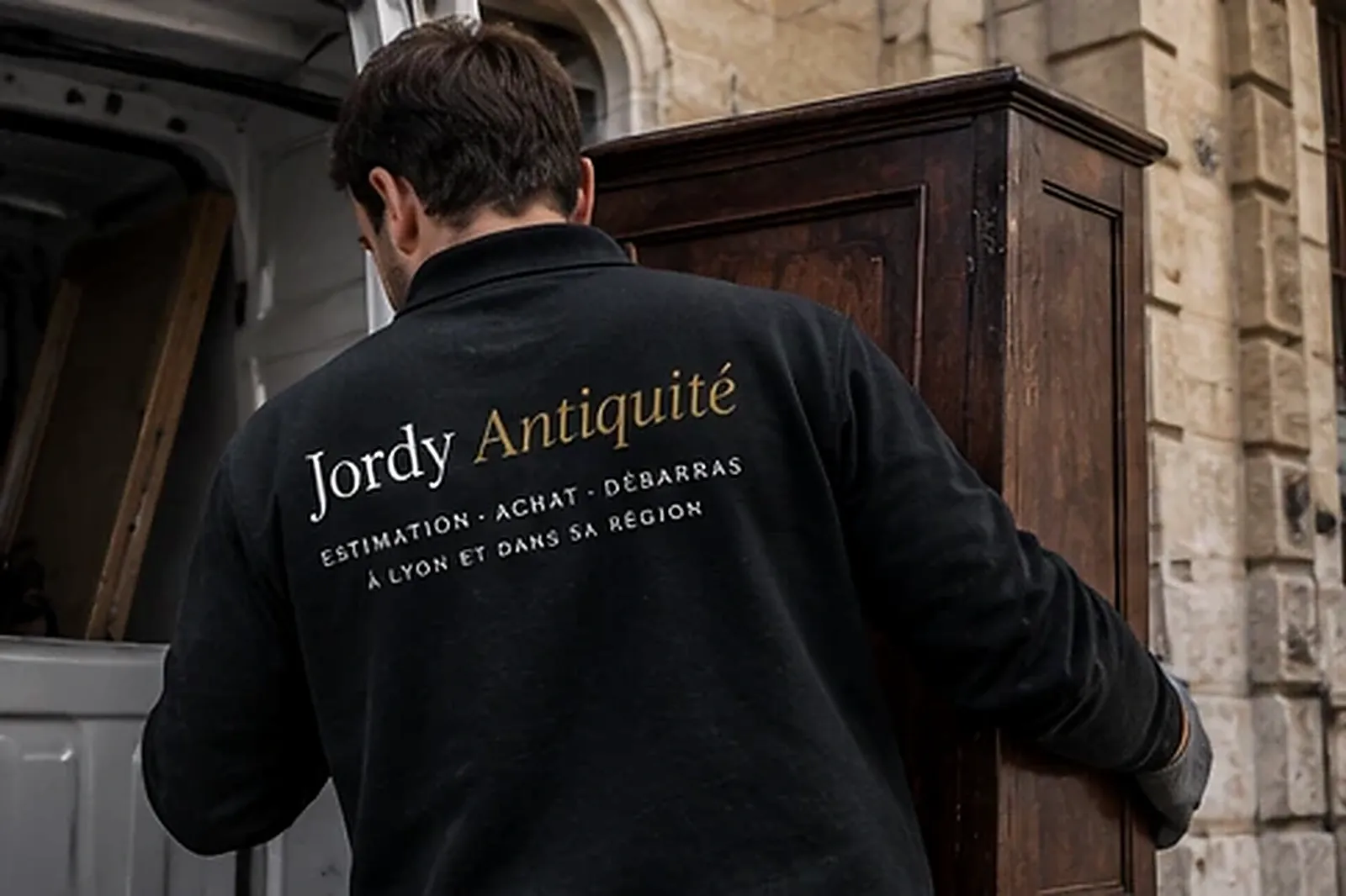 Un déménageur vu de dos, en polo noir floqué Jordy Dellinger, porte une grande armoire ancienne en bois sombre