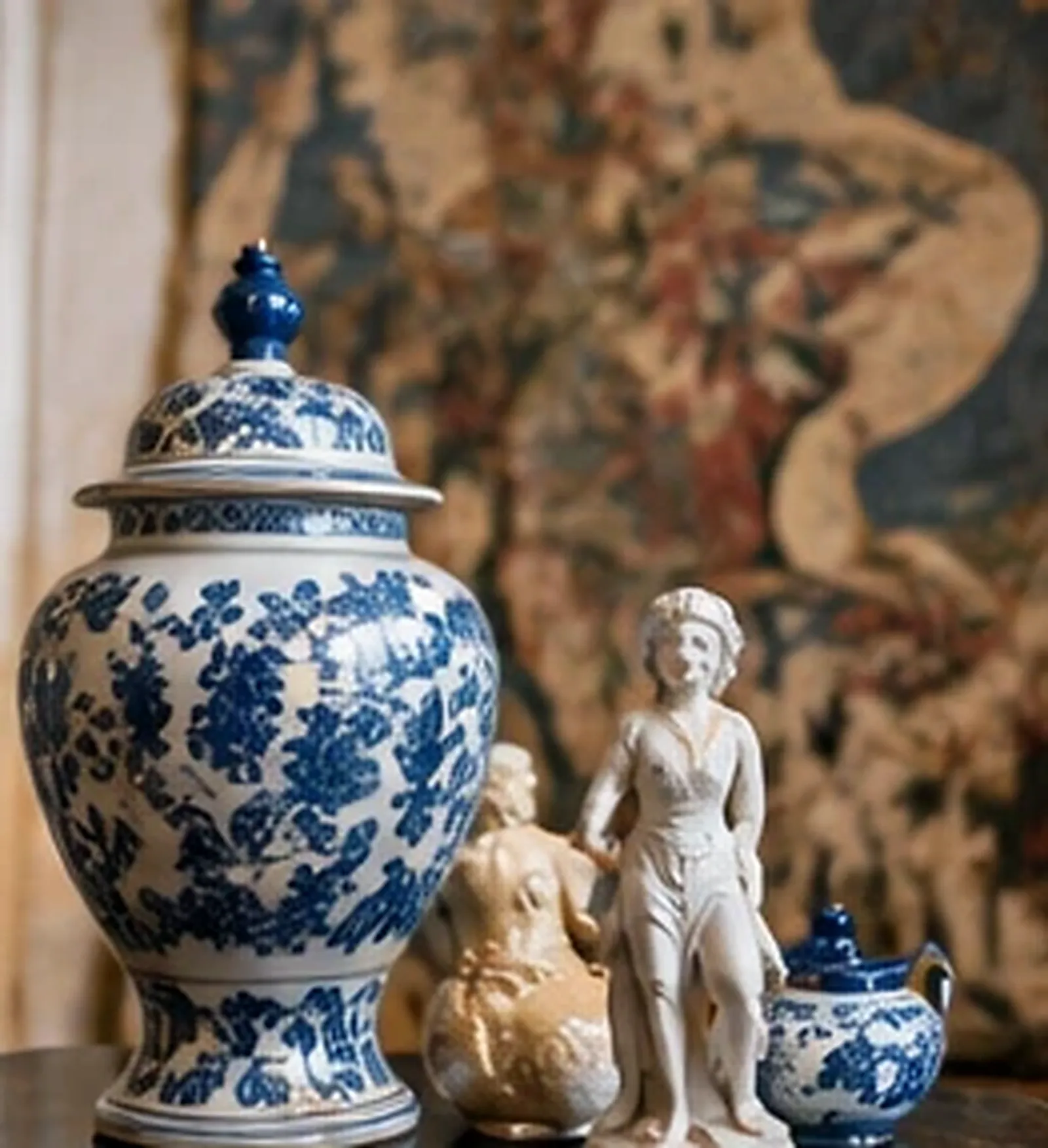 Potiche ancienne en porcelaine bleue et blanche à côté d'une statuette en biscuit et d'une verseuse, devant une tapisserie